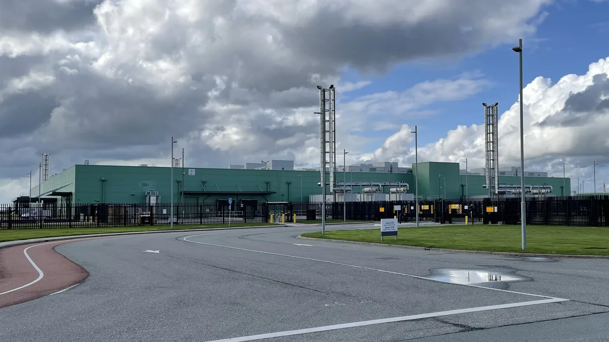 A large green data center building behind a black security fence under a cloudy blue sky.