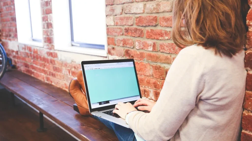 A woman sitting on a wooden bench against a brick wall, typing on a laptop with a green screen.