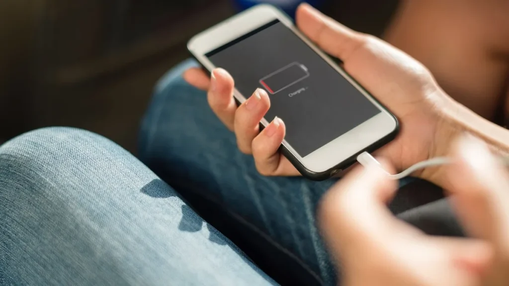 Hands holding a smartphone displaying a low red battery charging icon while plugged into a white cable.