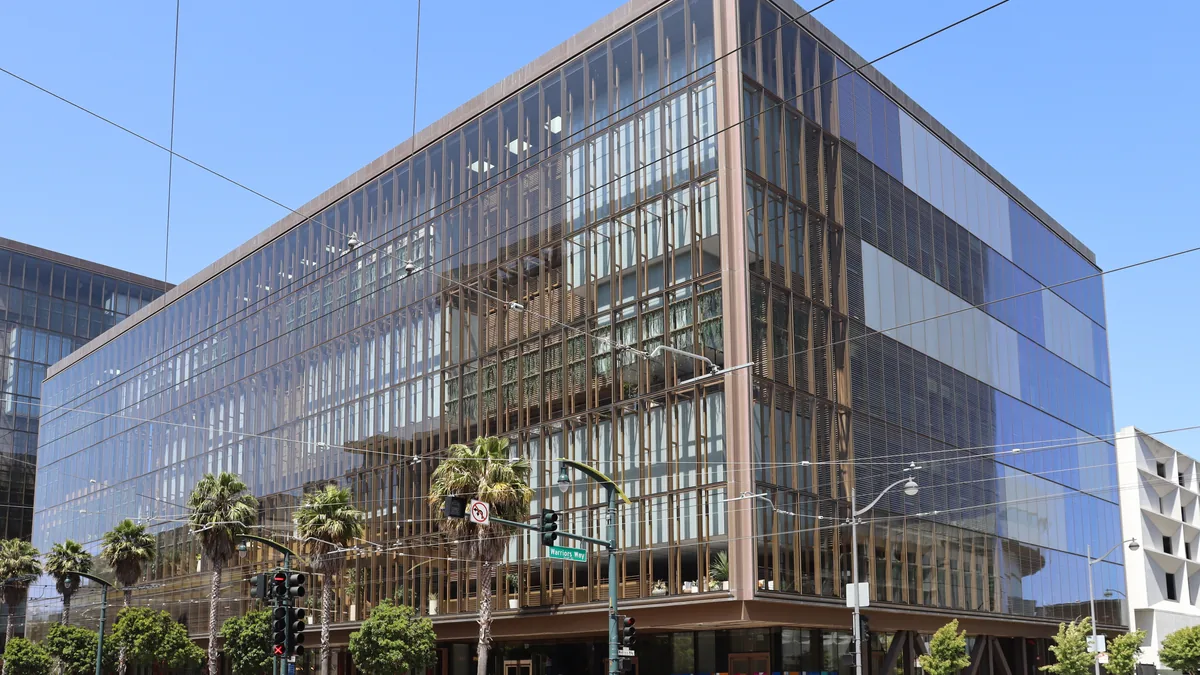 A modern glass office building with vertical columns stands behind palm trees under a clear blue sky.