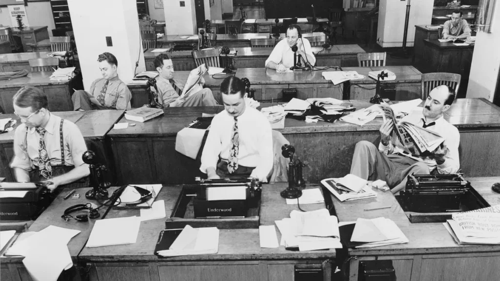 Vintage black and white photo of journalists working in a busy newsroom with typewriters and newspapers.