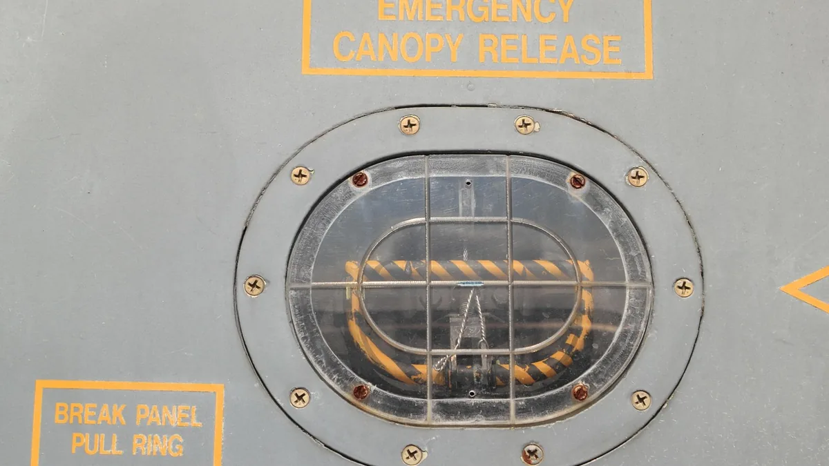 Emergency canopy release pull ring behind a protective grid on a grey metal panel with yellow labels.