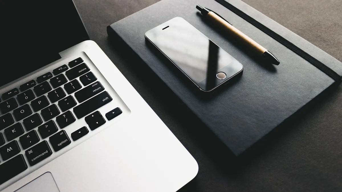 Laptop keyboard, smartphone, and pen resting on a black notebook in a dark, minimalist workspace.