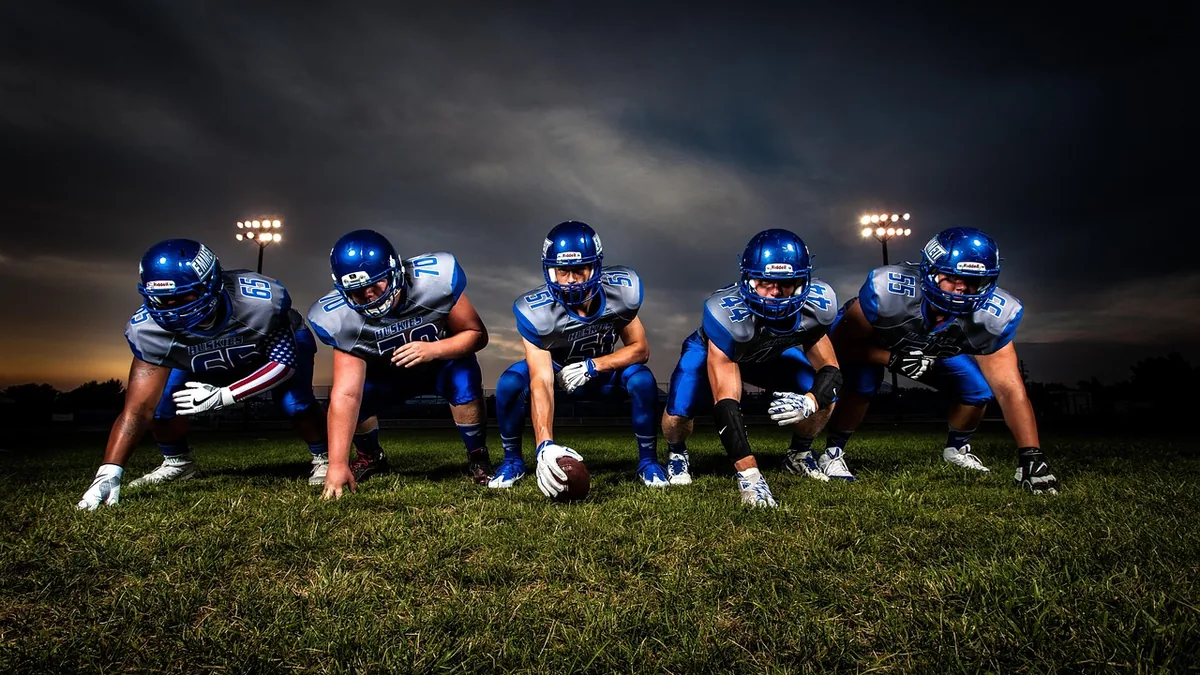 Five football players in blue uniforms crouching in a line on a grassy field under dramatic lighting.
