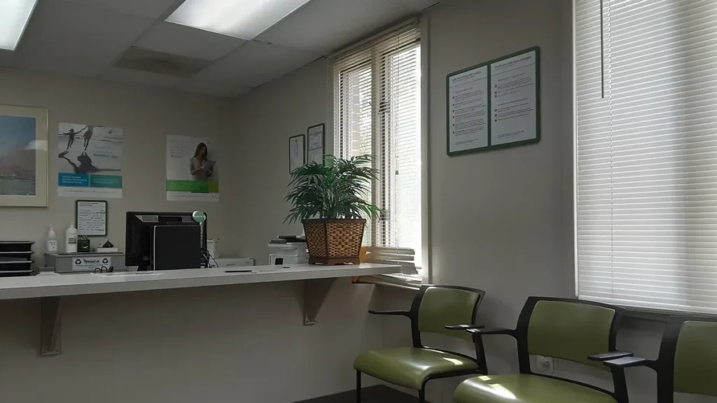 Reception desk in a medical clinic with green chairs, a potted plant, and computer monitor.
