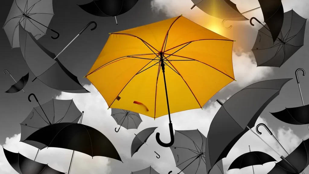 Bright yellow umbrella contrasting with falling black umbrellas under a cloudy sky.