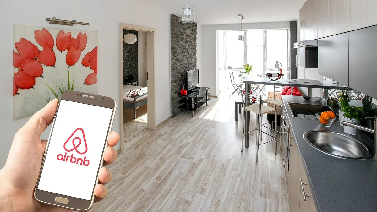 A hand holding a smartphone displaying the Airbnb logo in front of a modern apartment interior.