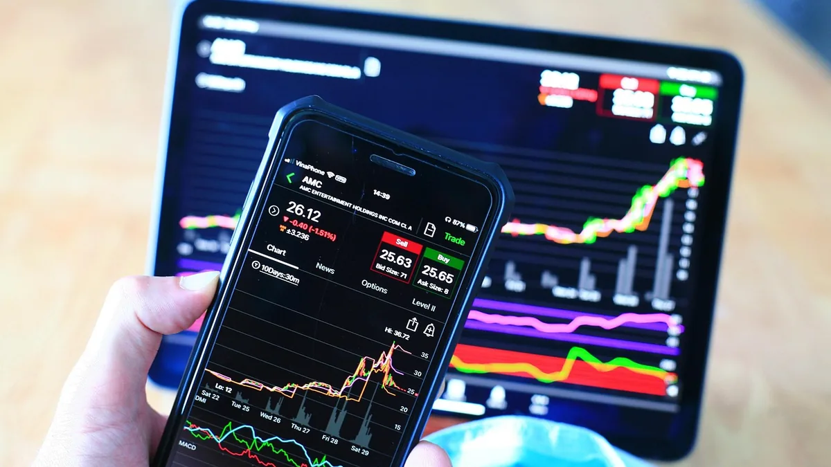 A hand holding a smartphone showing stock trading charts, with a tablet displaying financial graphs in the background.