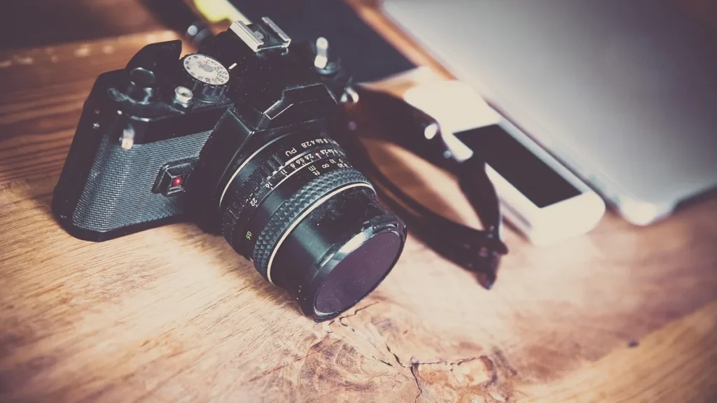 A vintage SLR camera, smartphone, and sunglasses resting on a wooden table.