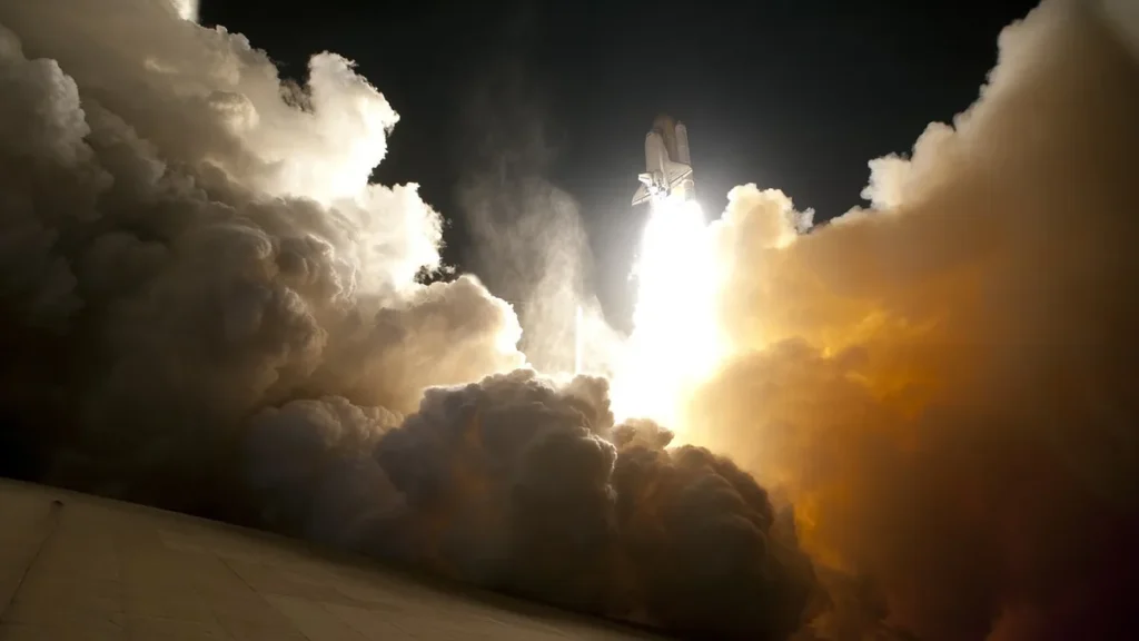 Space shuttle launching into the night sky, surrounded by huge clouds of bright fire and smoke.