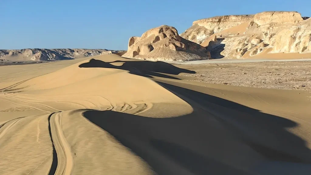 Sand dunes with tire tracks stretch towards rocky cliffs under a clear blue sky in a vast desert.