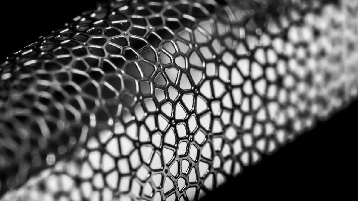 Black and white close-up of a metallic cylindrical mesh lattice structure against a dark background.
