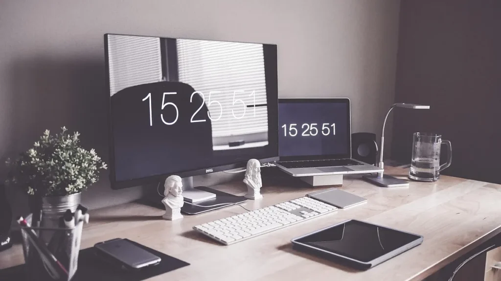A clean wooden desk setup with a large monitor, laptop displaying digital clocks, keyboard, tablet, and small plant.