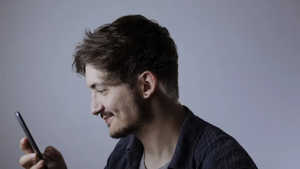 Profile of a young man smiling while looking at his smartphone against a plain background.