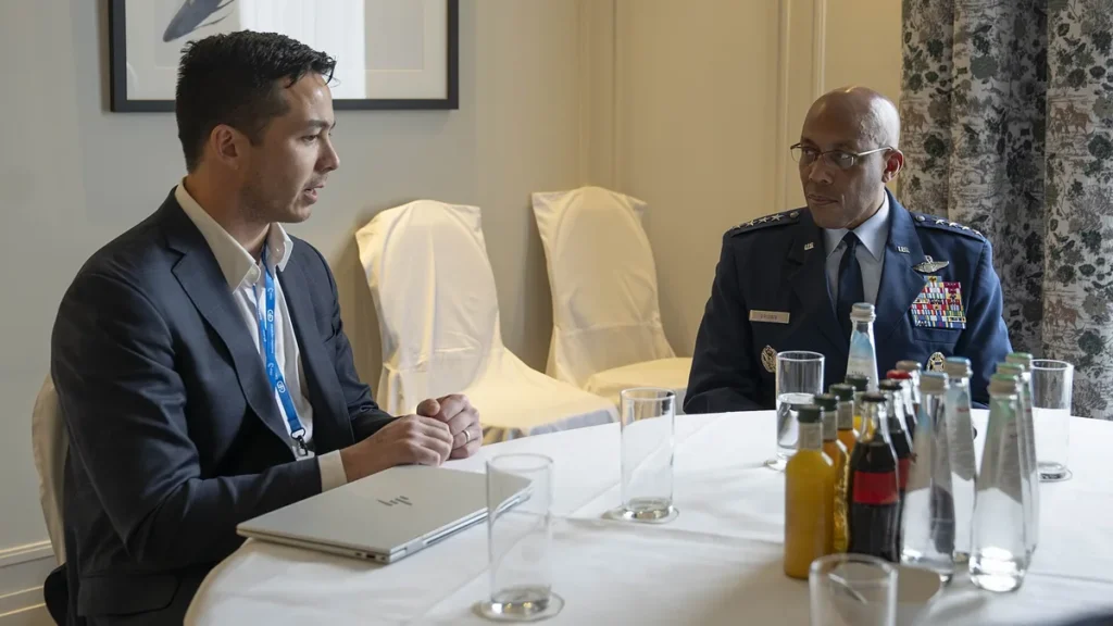 A civilian in a suit speaks with a US military general at a table with water bottles.