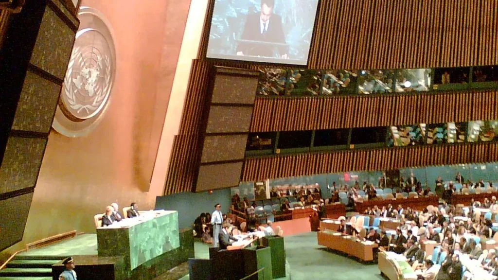 UN General Assembly hall showing the iconic emblem, delegates, and a speaker on the large overhead screen.