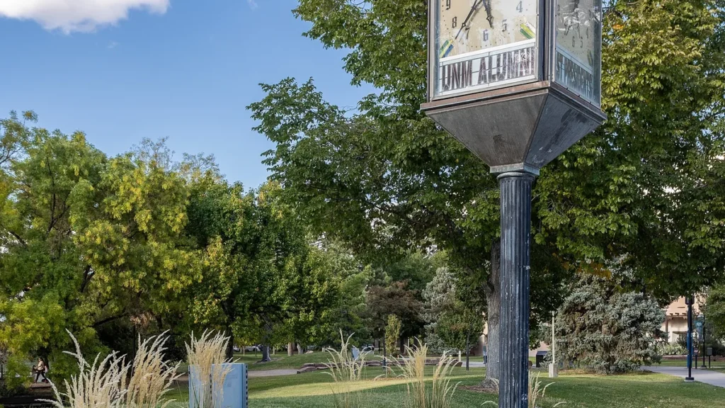 The UNM Alumni clock stands on a green, tree-lined campus under a blue sky.