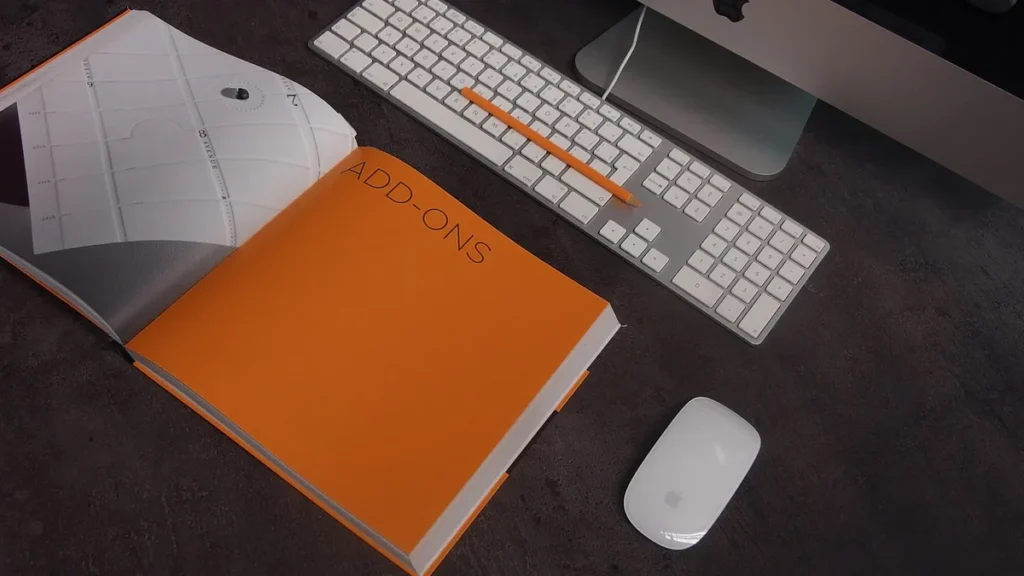 Orange book page 'ADD-ONS' beside white keyboard, orange pencil, and mouse on dark desk.