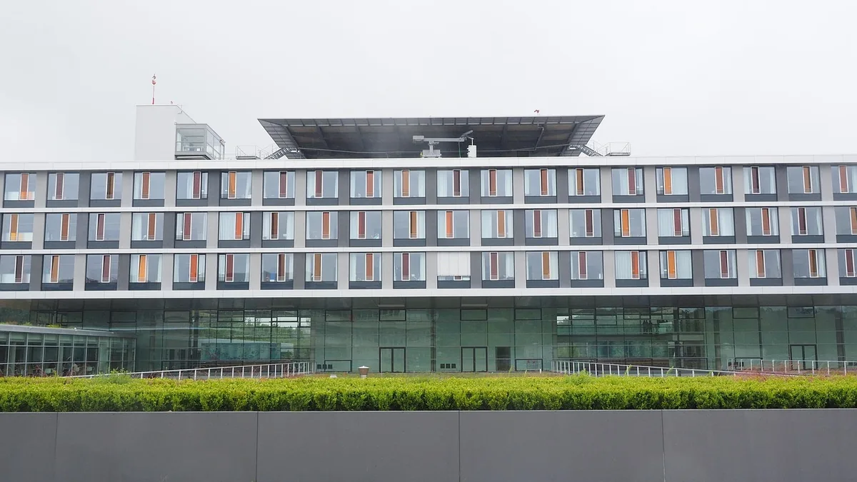 Modern hospital building with a rooftop helipad, viewed from behind a green hedge under a cloudy sky.