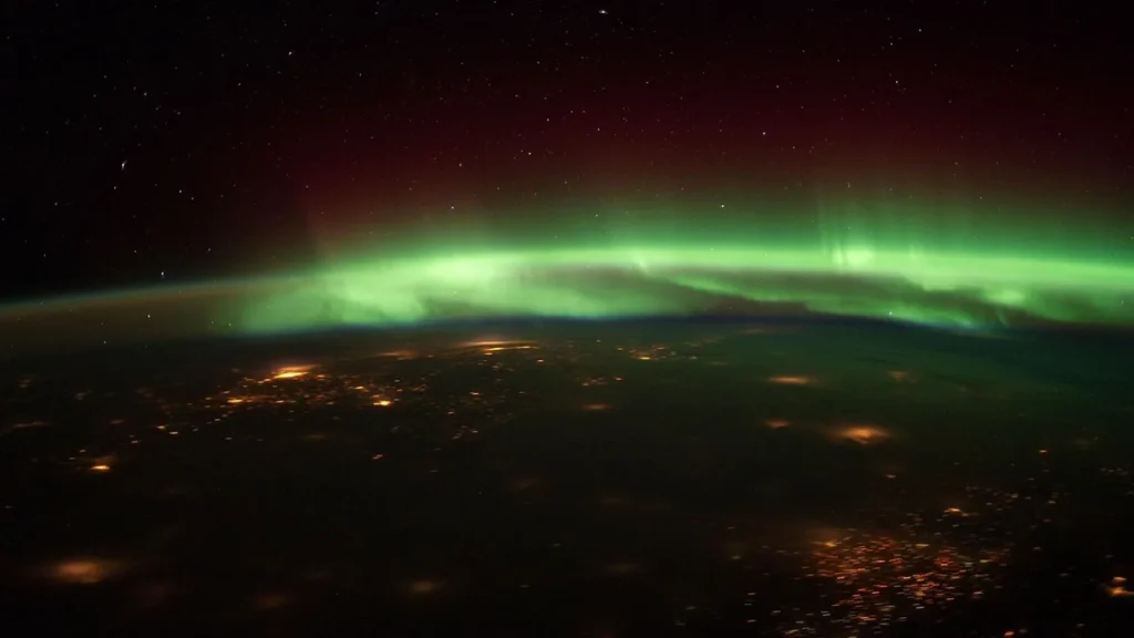 Vibrant green aurora borealis stretches over Earth's horizon, dotted with glowing city lights under a starry sky.
