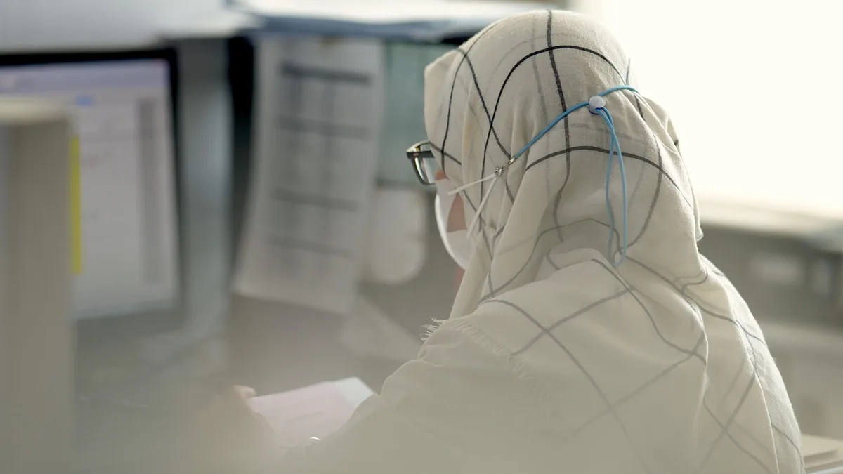 Person wearing a hijab and face mask, viewed from behind while working at a computer.