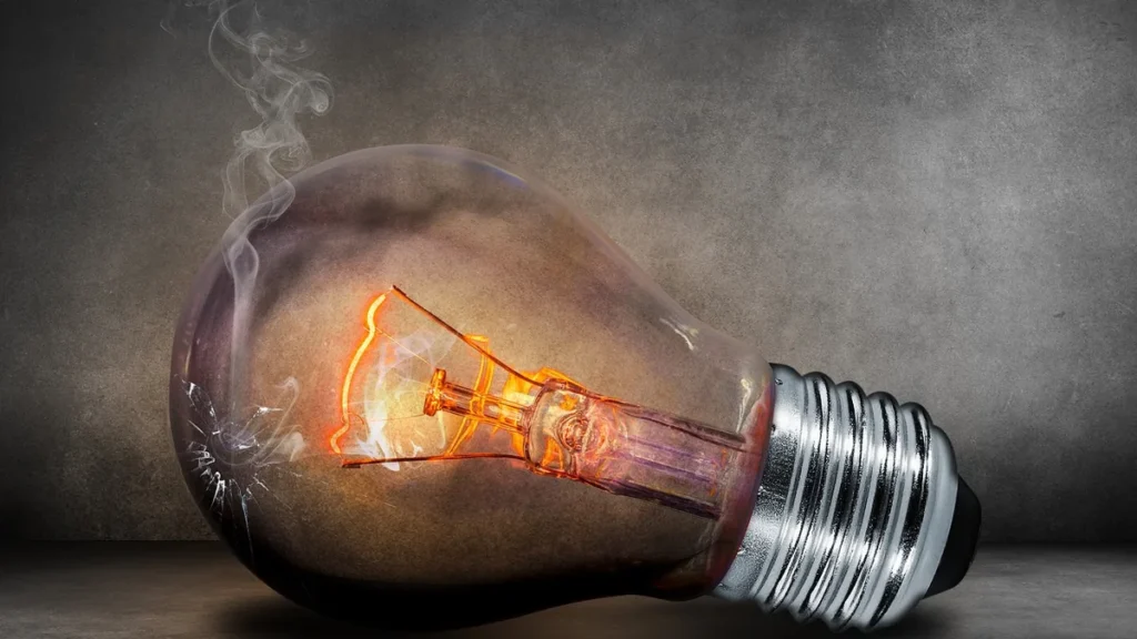 A cracked, smoking light bulb with a glowing orange filament lies on a dark grey surface.