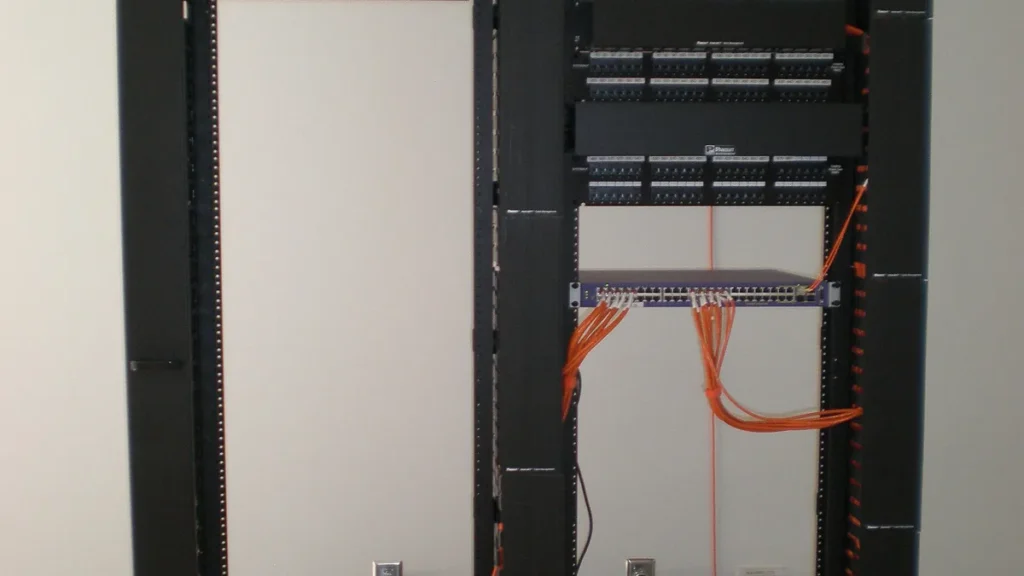 A server rack showing patch panels and a network switch with orange ethernet cables.