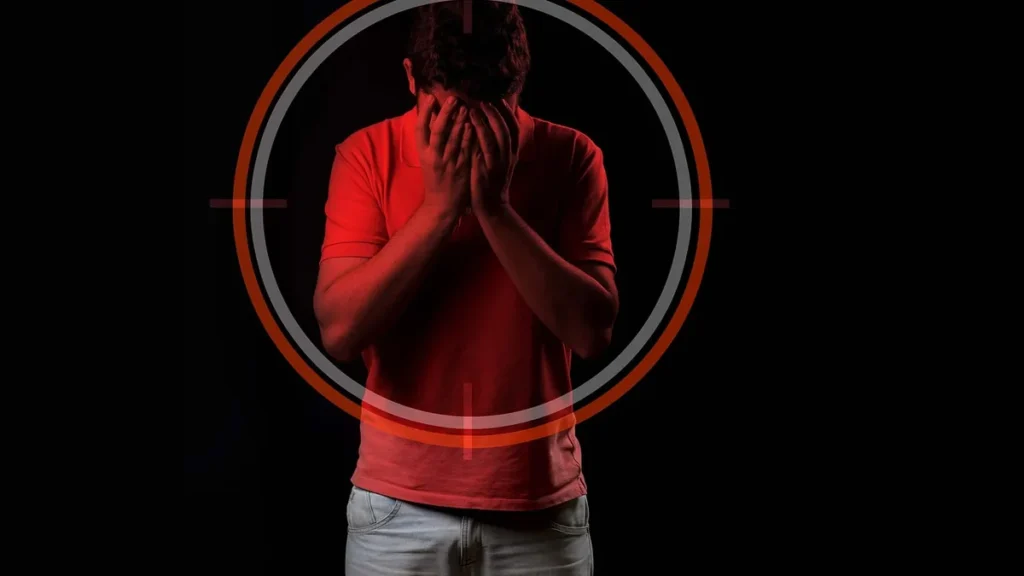 Man covering his face under a red target crosshair on a black background.