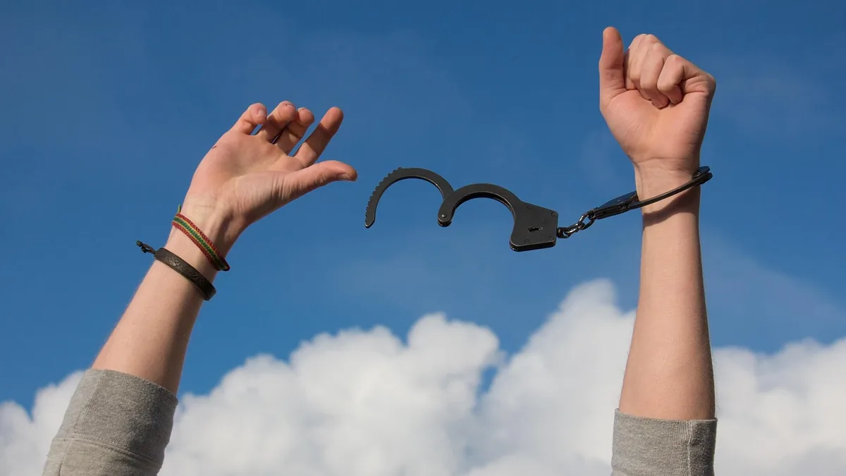Two arms raised against a blue sky with clouds; one wrist is in a broken handcuff.
